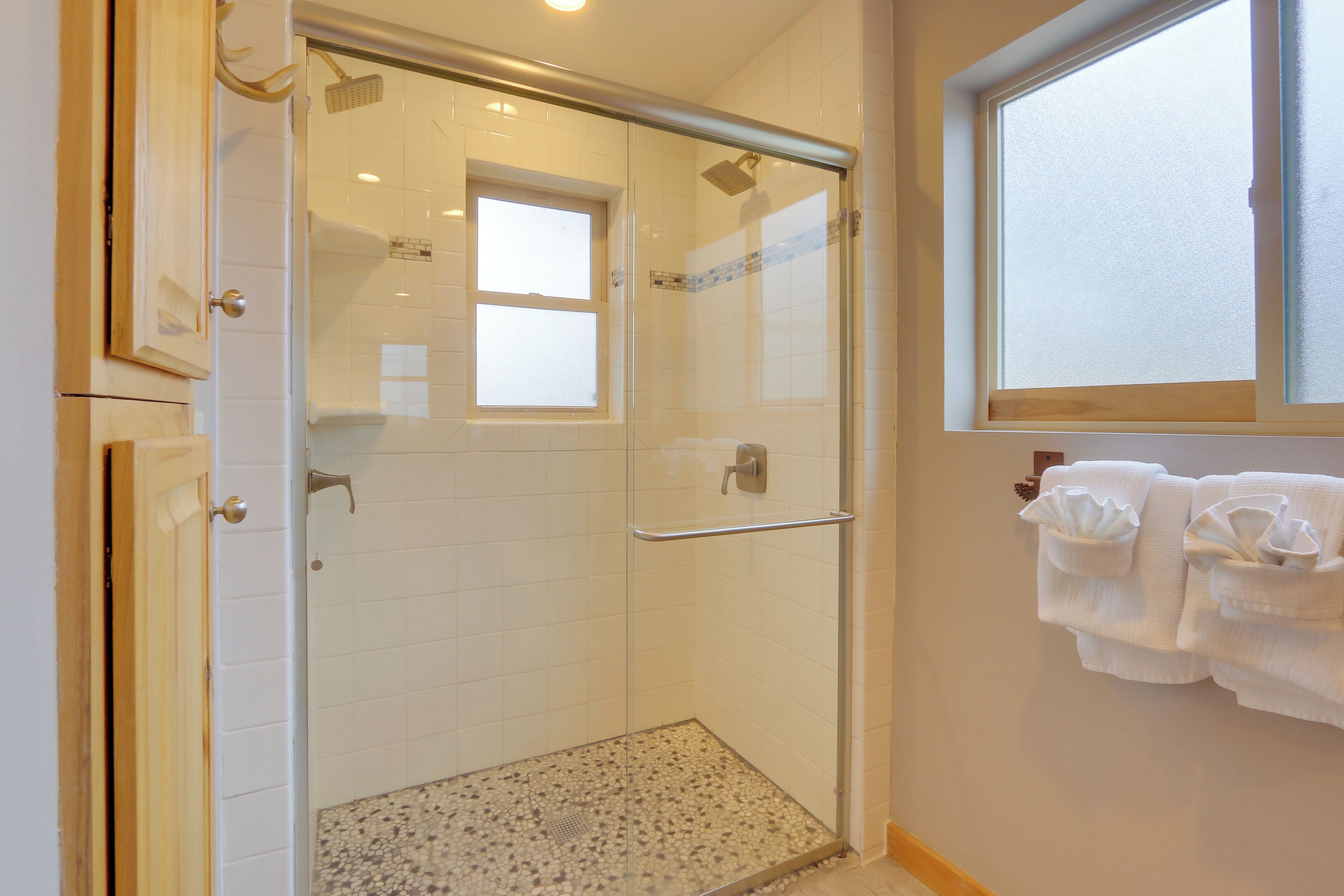 Ducha con mampara de vidrio, una ventana pequeña, un perchero con toallas y una puerta de madera.