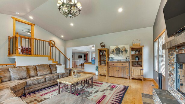 Un salon spacieux avec un grand canapé d’angle, une table basse en bois et un tapis à motifs. Un escalier avec une rampe en bois mène à l’étage supérieur. Une cheminée avec un manteau en pierre est visible sur le côté droit.