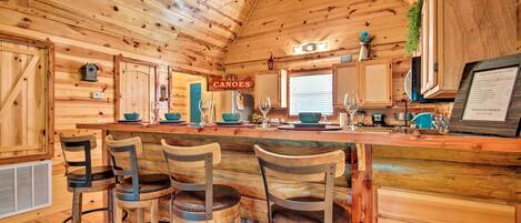 A wooden cabin interior with a bar area, four barstools, a refrigerator, and a sign indicating 'Canoes' on the wall.