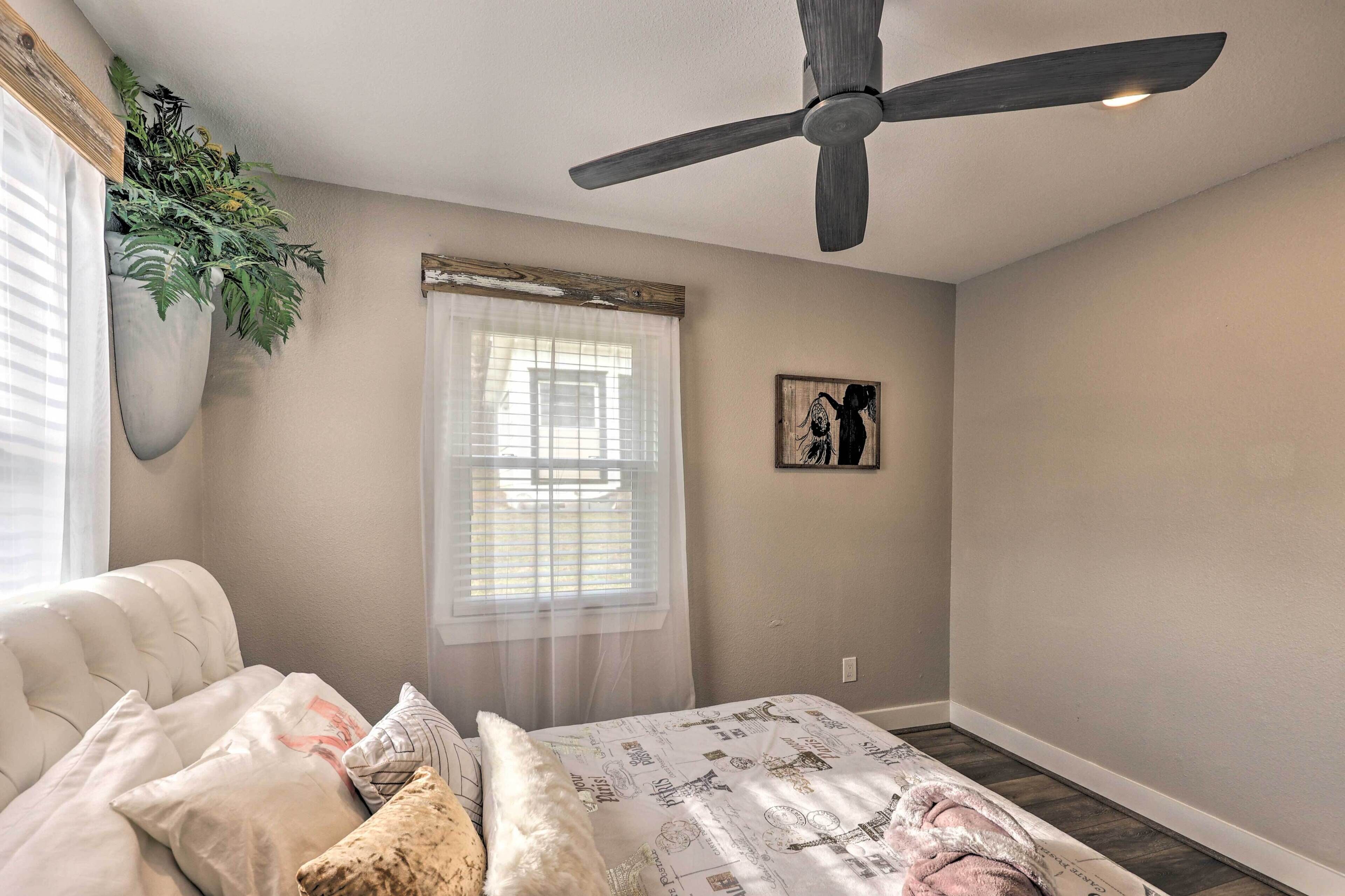 A bedroom with a bed, a ceiling fan, a window with sheer curtains, and a wall-mounted plant.