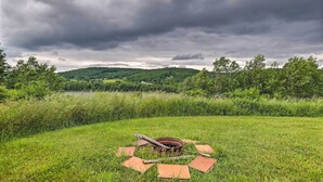 Un espace extérieur rustique, avec un foyer en métal entouré de briques, le tout se fondant dans un décor de verdure luxuriante et surplombant un lac sous un ciel nuageux.