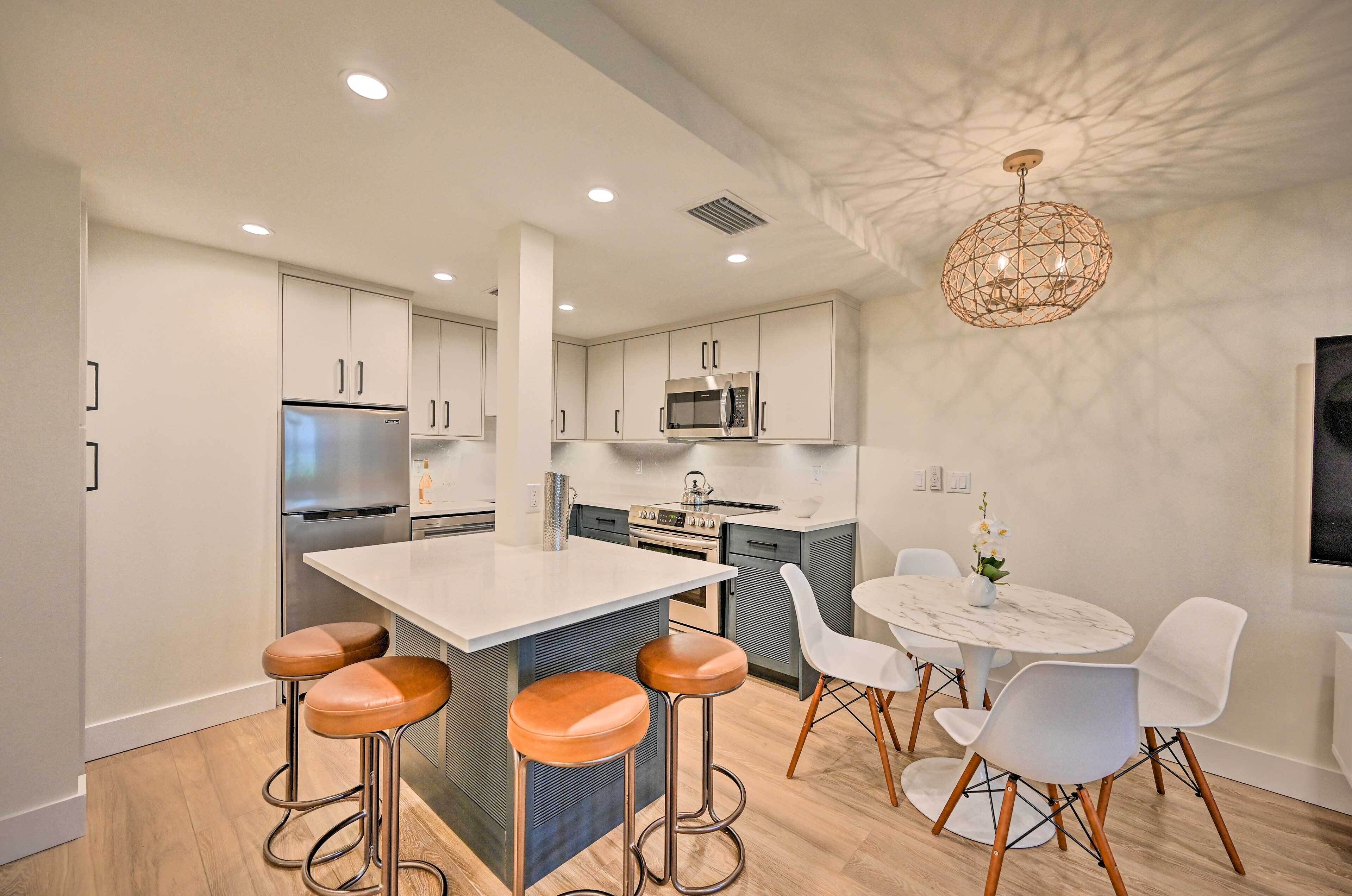 A modern kitchen with a central island, bar stools, a dining table with chairs, and a unique ceiling light fixture.