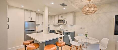 A modern kitchen with a central island, bar stools, a dining table with chairs, and a unique ceiling light fixture.