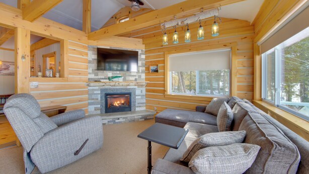 A log cabin interior with a stone fireplace, wooden beams, a grey sofa, and a window with a view of trees. - Palmyra Cottage w/ Deck on Whites Pond! (Palmyra)