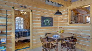 A cozy cabin interior with a bed, a dining table with chairs, a chandelier, and a framed sign on the wall. - Palmyra Cottage w/ Deck on Whites Pond! (Palmyra)
