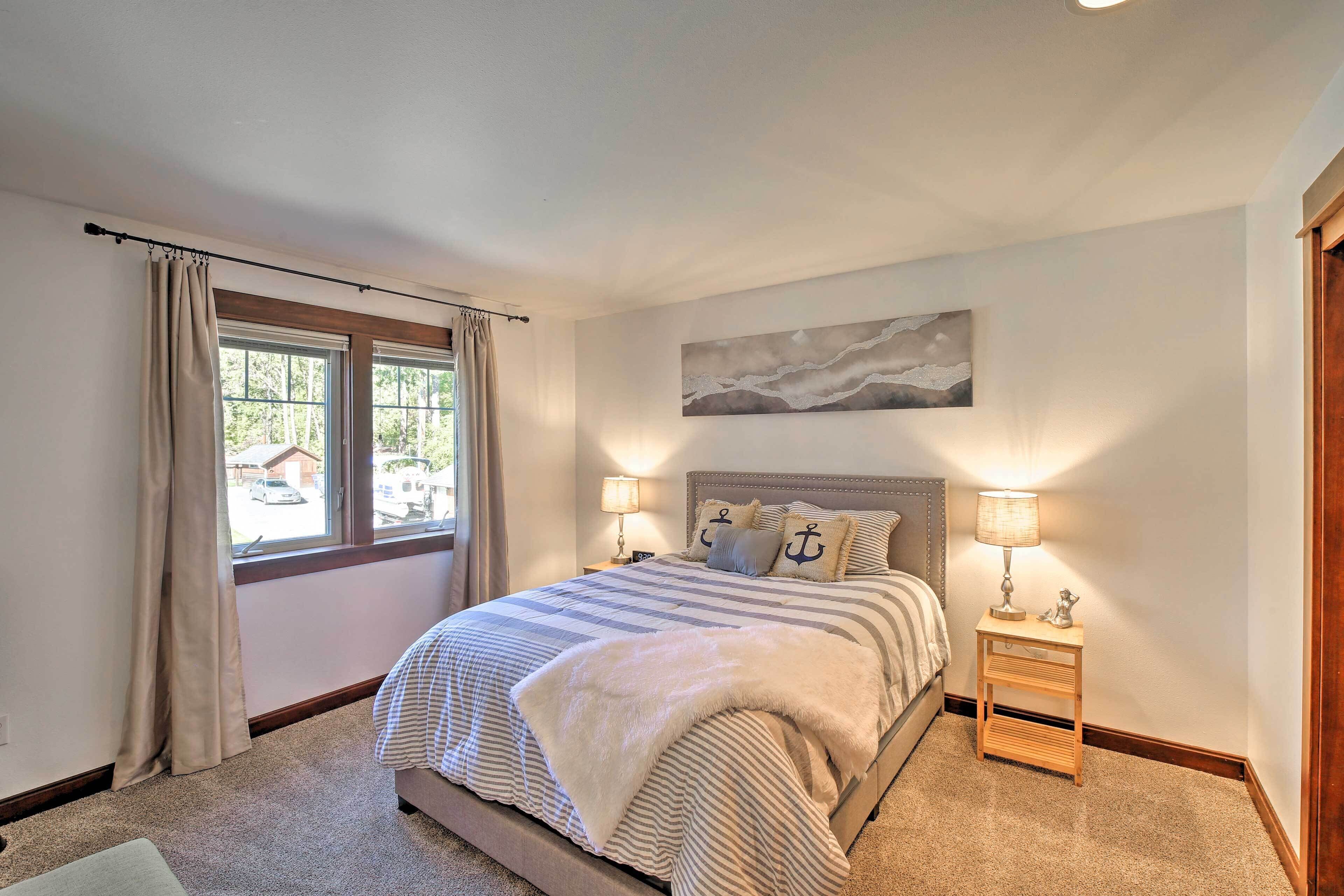 A bedroom with a large bed, two bedside lamps, a window with curtains, and a painting on the wall.