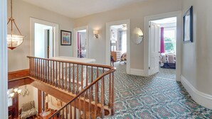 A spacious hallway with a wooden staircase, patterned carpet, and a view into a bedroom with a bed and window. - Spacious Victorian Home 1/4 Mi to Holiday Valley! (Ellicottville)