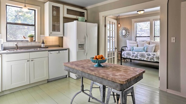 A kitchen with white cabinets, a white refrigerator, a wooden table with a bowl of fruit, and a window with a view of the outdoors.