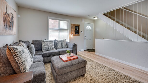 Een woonkamer met een grijze hoekbank, een houten salontafel en een beige vloerkleed. Er is een trap met witte leuningen die naar een verdieping leidt.