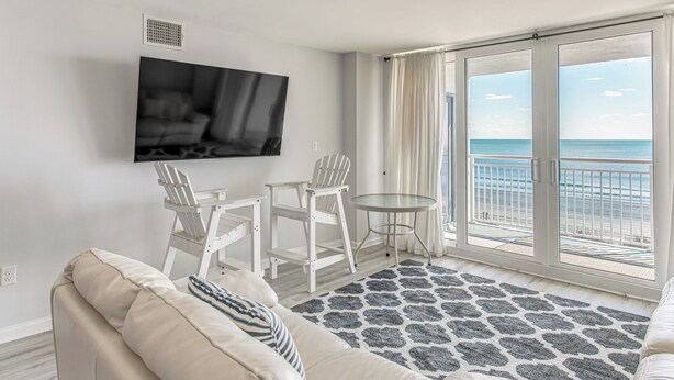 A modern living room with a large TV, white furniture, a balcony with a view of the sea, and a patterned rug.