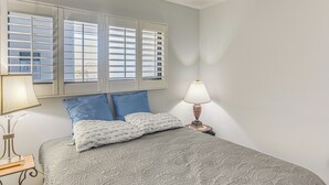 A bedroom with a bed, two pillows, a nightstand with a lamp, and a window with blinds.