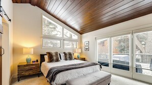 A bedroom with a large bed, two bedside tables with lamps, a bench, and a view of the outside through large windows. - Modern Mountain Escape on Golf Course w/ Hot Tub! (Avon)