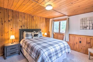 A bedroom with a wooden ceiling, a plaid bedspread, two bedside lamps, a nightstand with drawers, a window with curtains, and a wall-mounted painting.