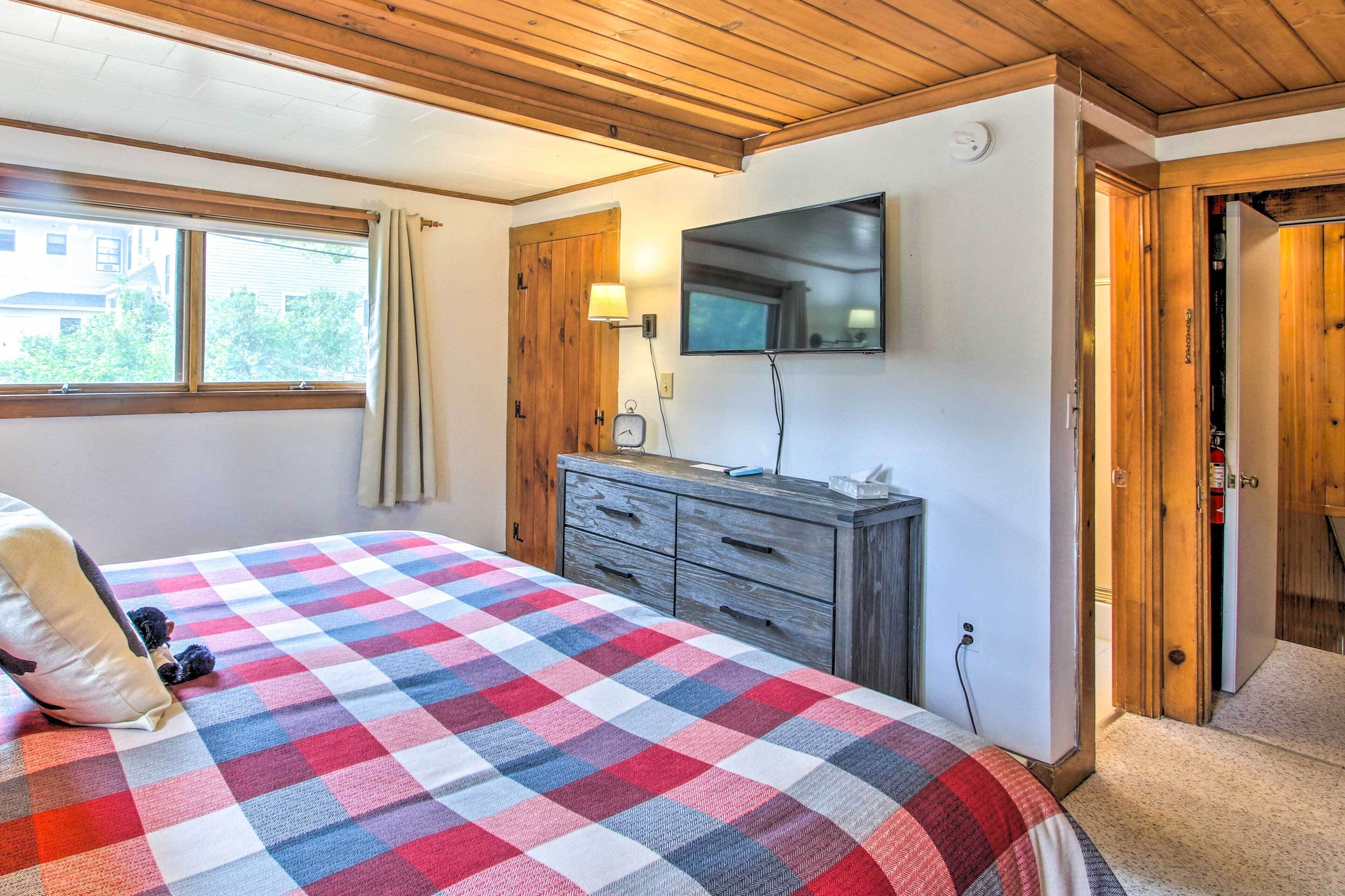 Un dormitorio con colcha a cuadros, cómoda de madera, televisor montado en la pared y ventana con cortinas.