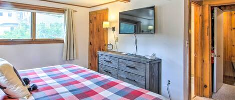 Un dormitorio con colcha a cuadros, cómoda de madera, televisor montado en la pared y ventana con cortinas.