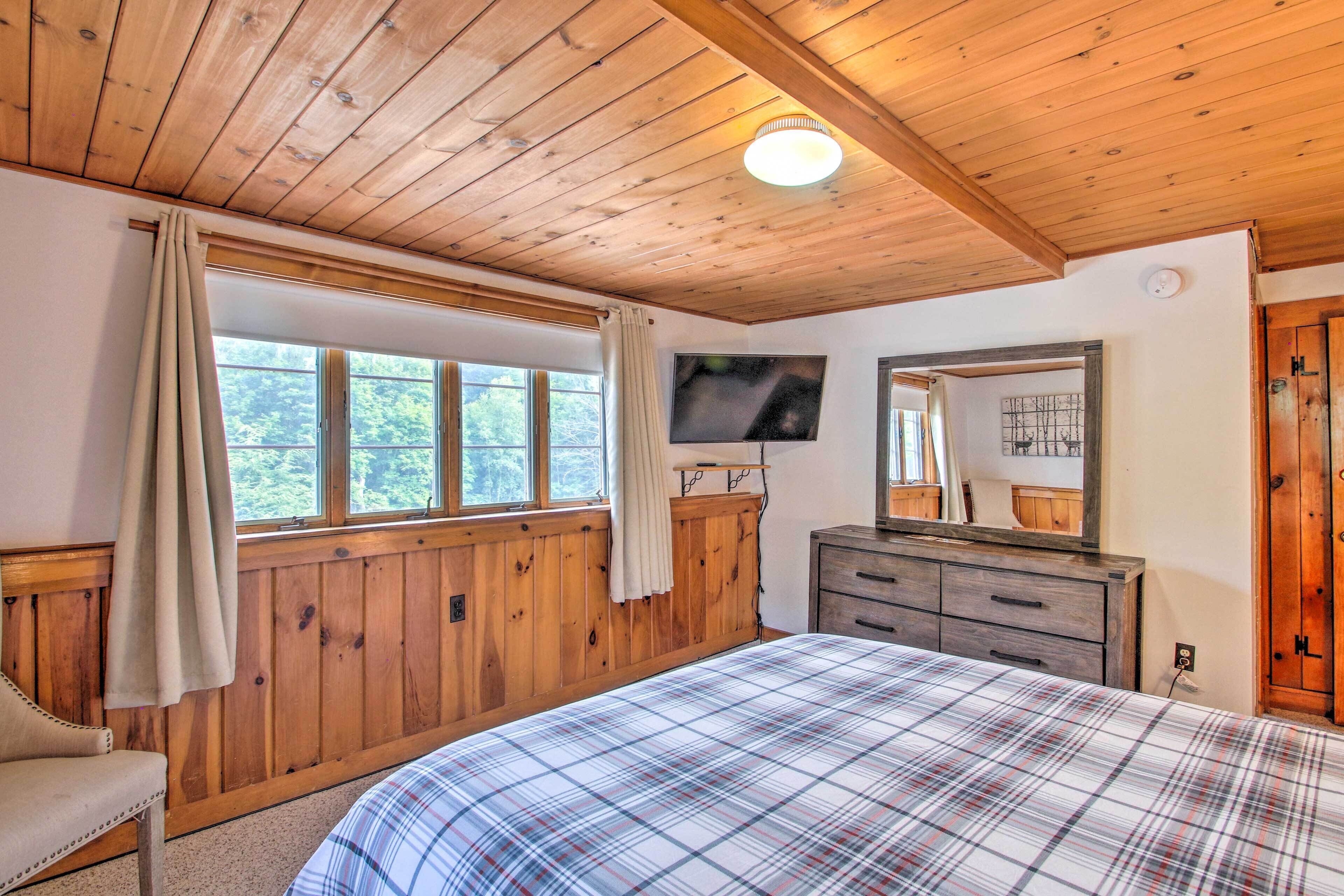 Une chambre avec des lambris en bois, un lit recouvert d’une couverture à carreaux, un téléviseur posé sur une commode et une fenêtre avec des rideaux.