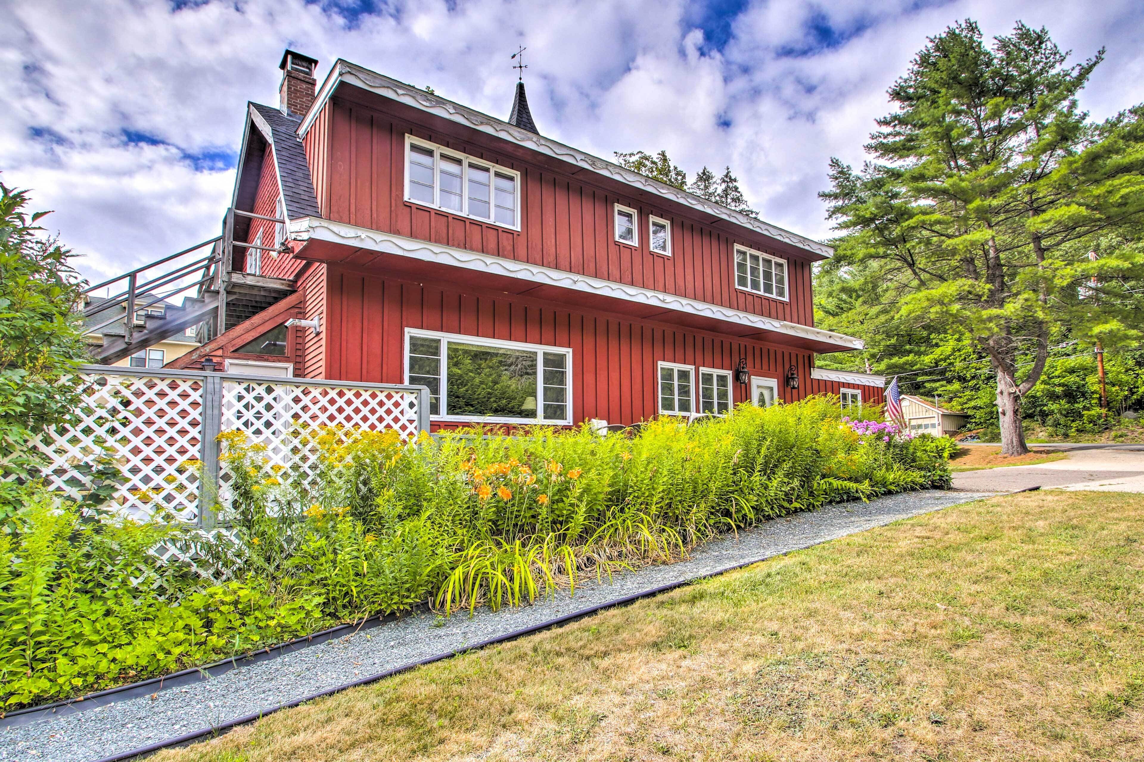 A red two-story house with a white fence, a small garden with various plants, and a clear sky.
