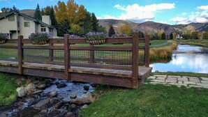 Un puente de madera sobre un arroyo, con macetas de flores en la barandilla, una casa al fondo y una cadena montañosa.
