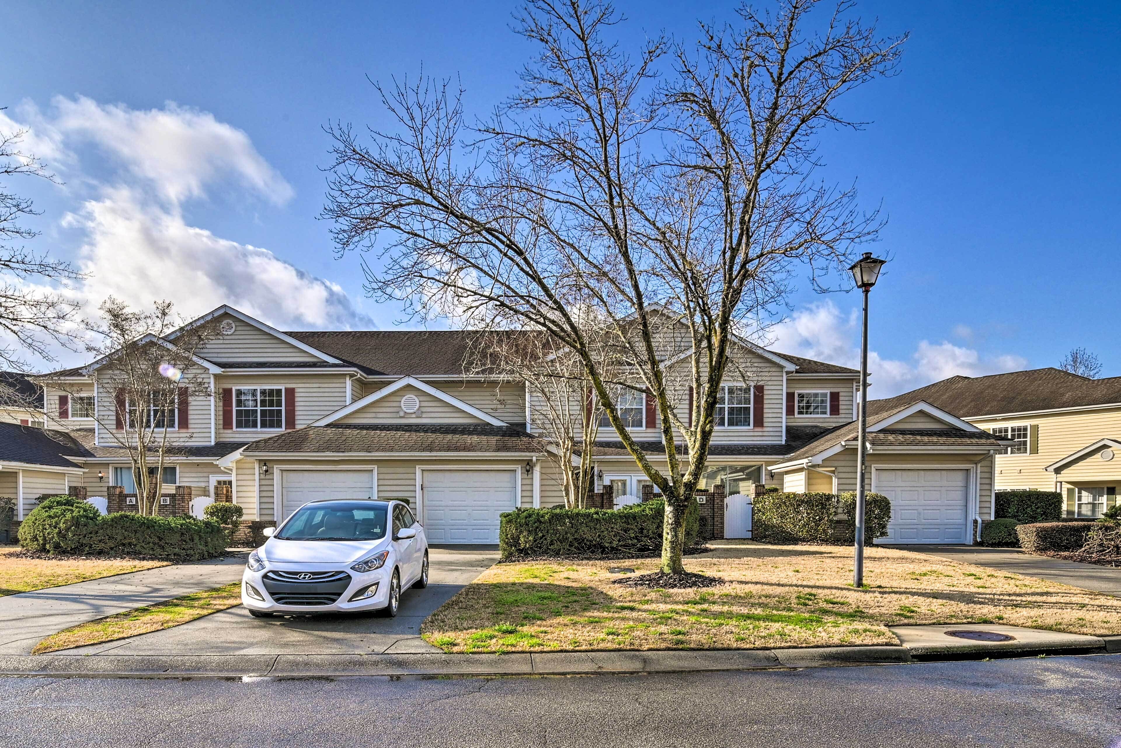 A residential street with houses, a white car parked in the driveway, and a clear blue sky.