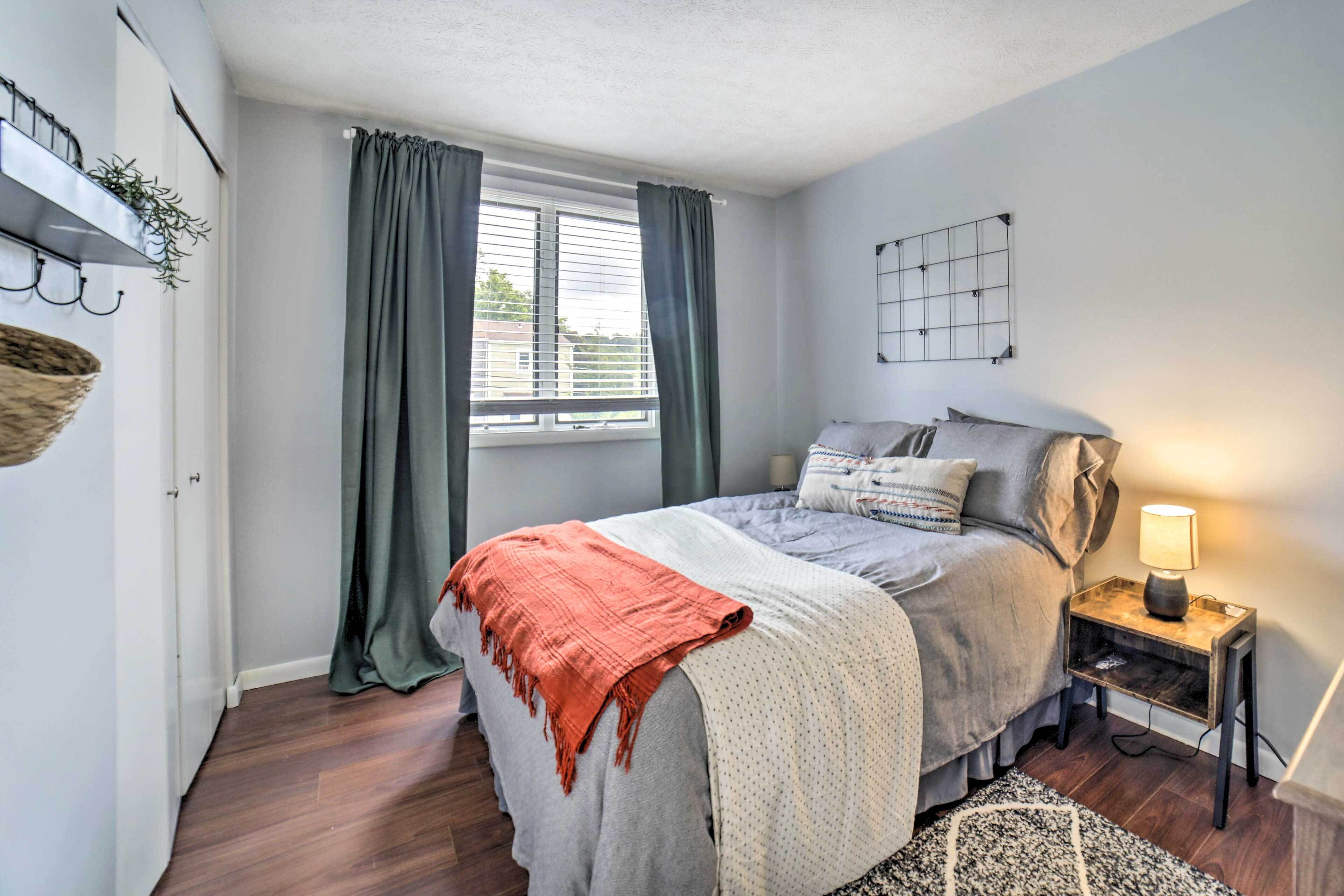 A bedroom with a bed, a bedside table with a lamp, a window with curtains, a wall-mounted shelf with plants, and a wooden floor.