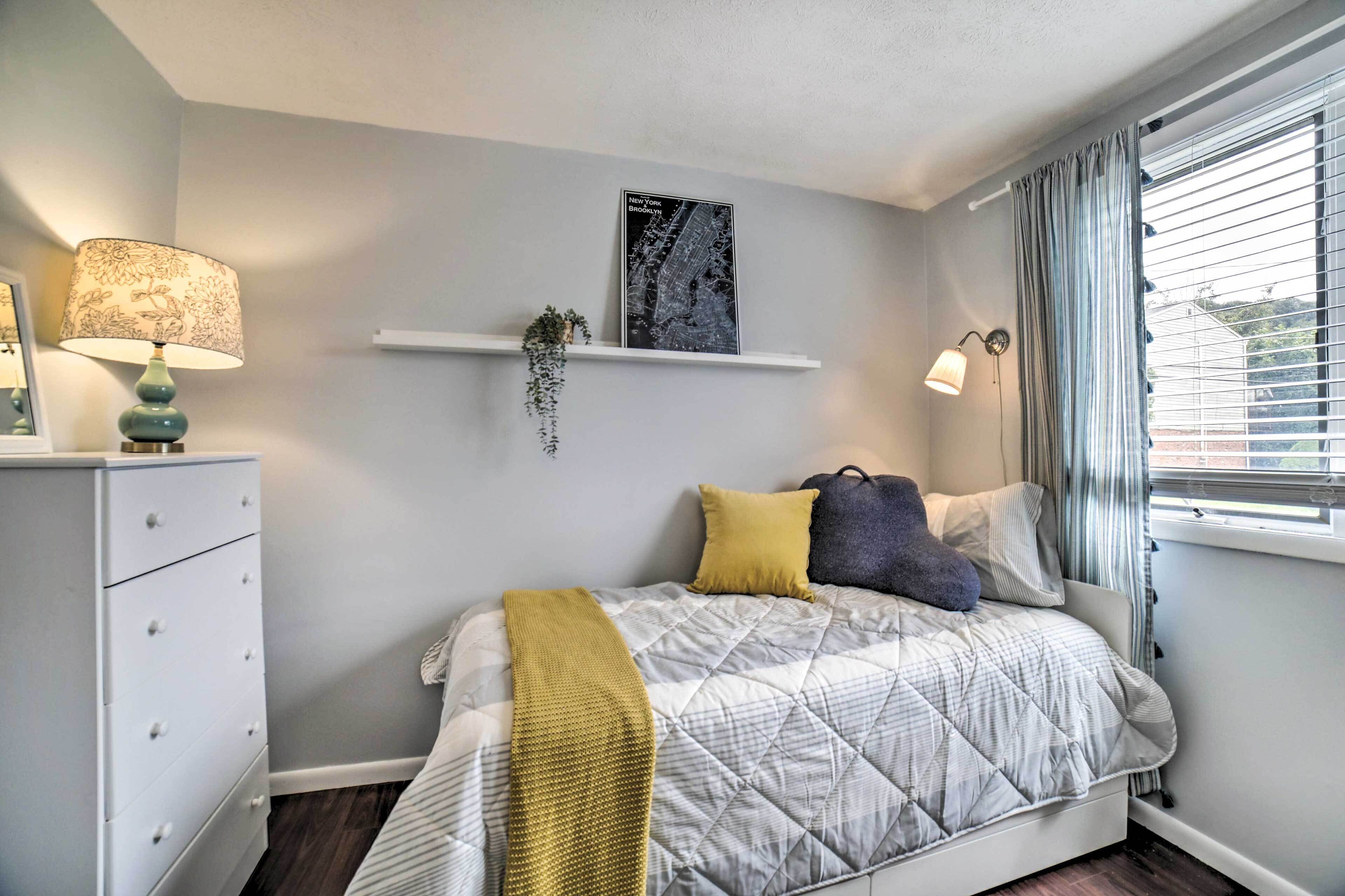 Un dormitorio con cama, mesita de noche, lámpara, estante con una planta, mapa enmarcado y ventana con cortinas.