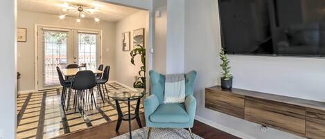 A living room with a TV, a teal armchair, a dining area with a table and chairs, and a patterned rug.