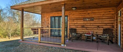 Une cabane en rondins dotée d’un porche couvert, de deux fauteuils Adirondack, d’une petite table et d’une fenêtre.