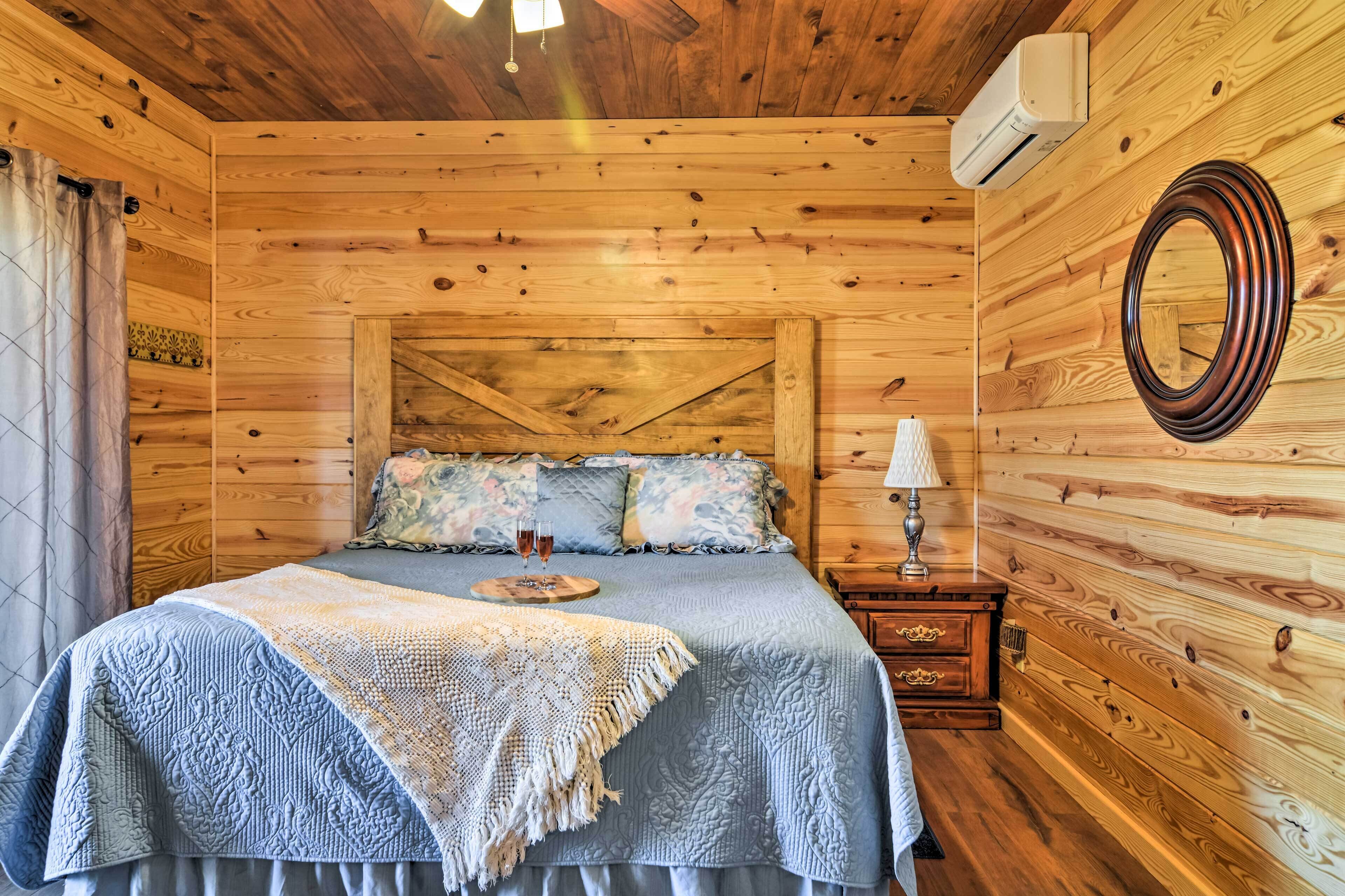 A wooden cabin bedroom with a bed, a nightstand, a mirror, and a window with curtains.