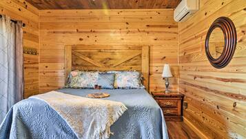 A wooden cabin bedroom with a bed, a nightstand, a mirror, and a window with curtains.