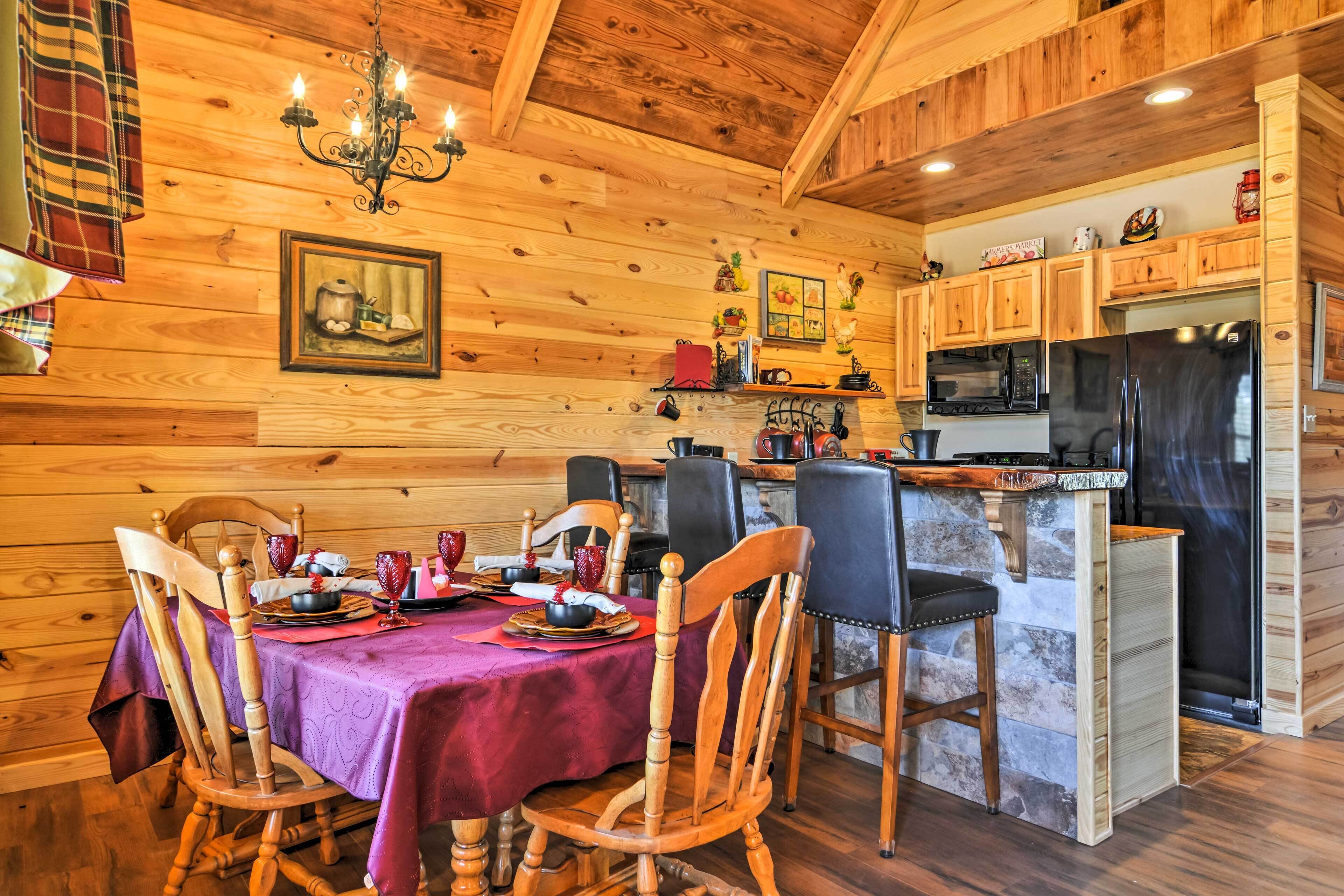 A wooden cabin interior with a dining table set for four, wooden chairs, a kitchen area with modern appliances, and a chandelier.