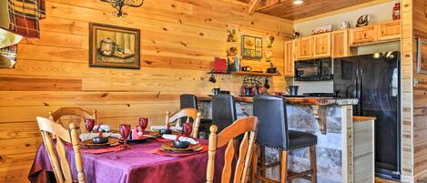 A wooden cabin interior with a dining table set for four, wooden chairs, a kitchen area with modern appliances, and a chandelier.