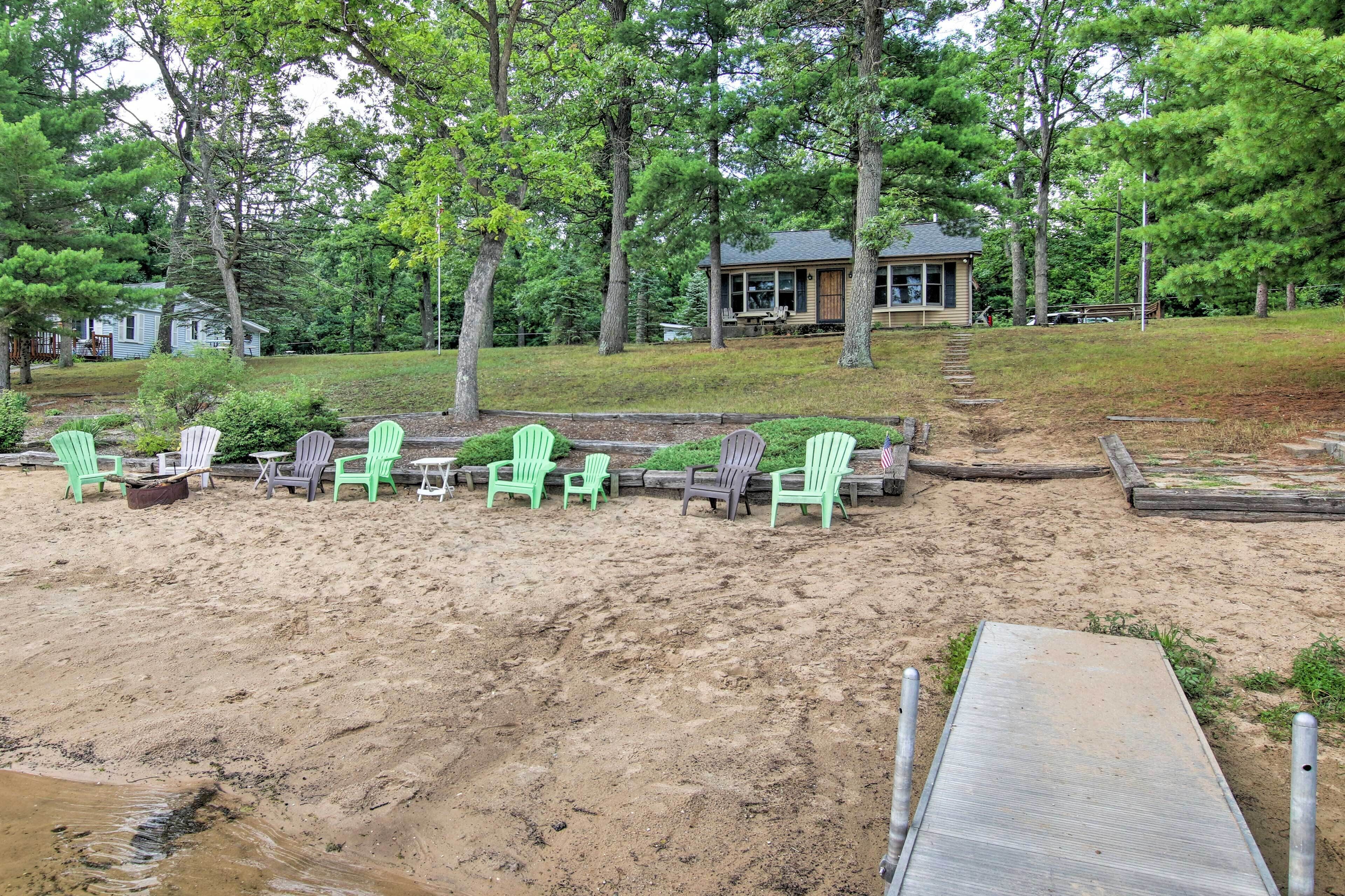 A sandy beach area with green Adirondack chairs, a wooden deck, and a house in the background.