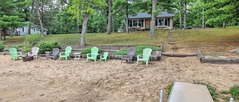 A sandy beach area with green Adirondack chairs, a wooden deck, and a house in the background.