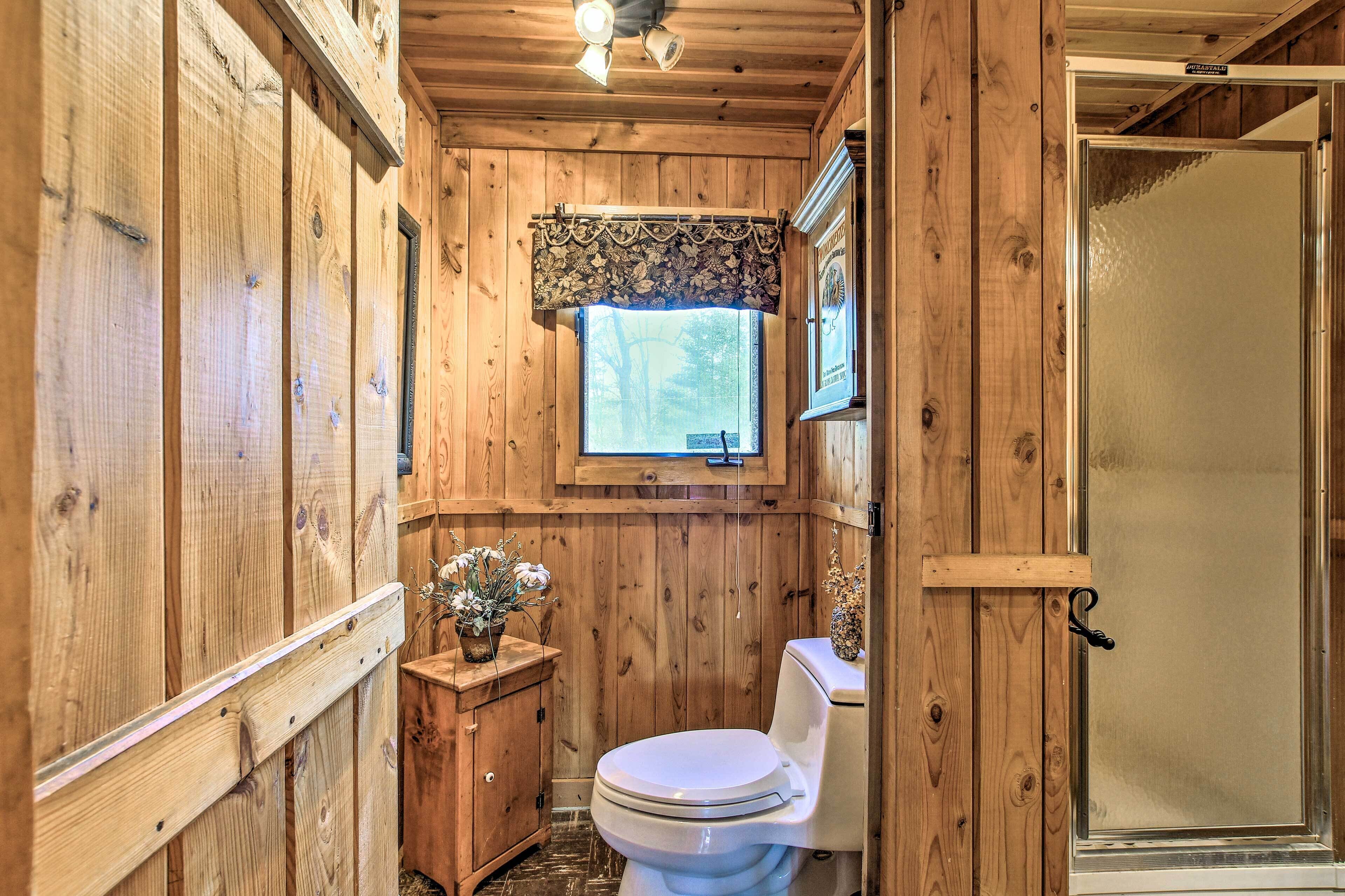 A wooden bathroom with a toilet, a small cabinet, and a window with a decorative valance.