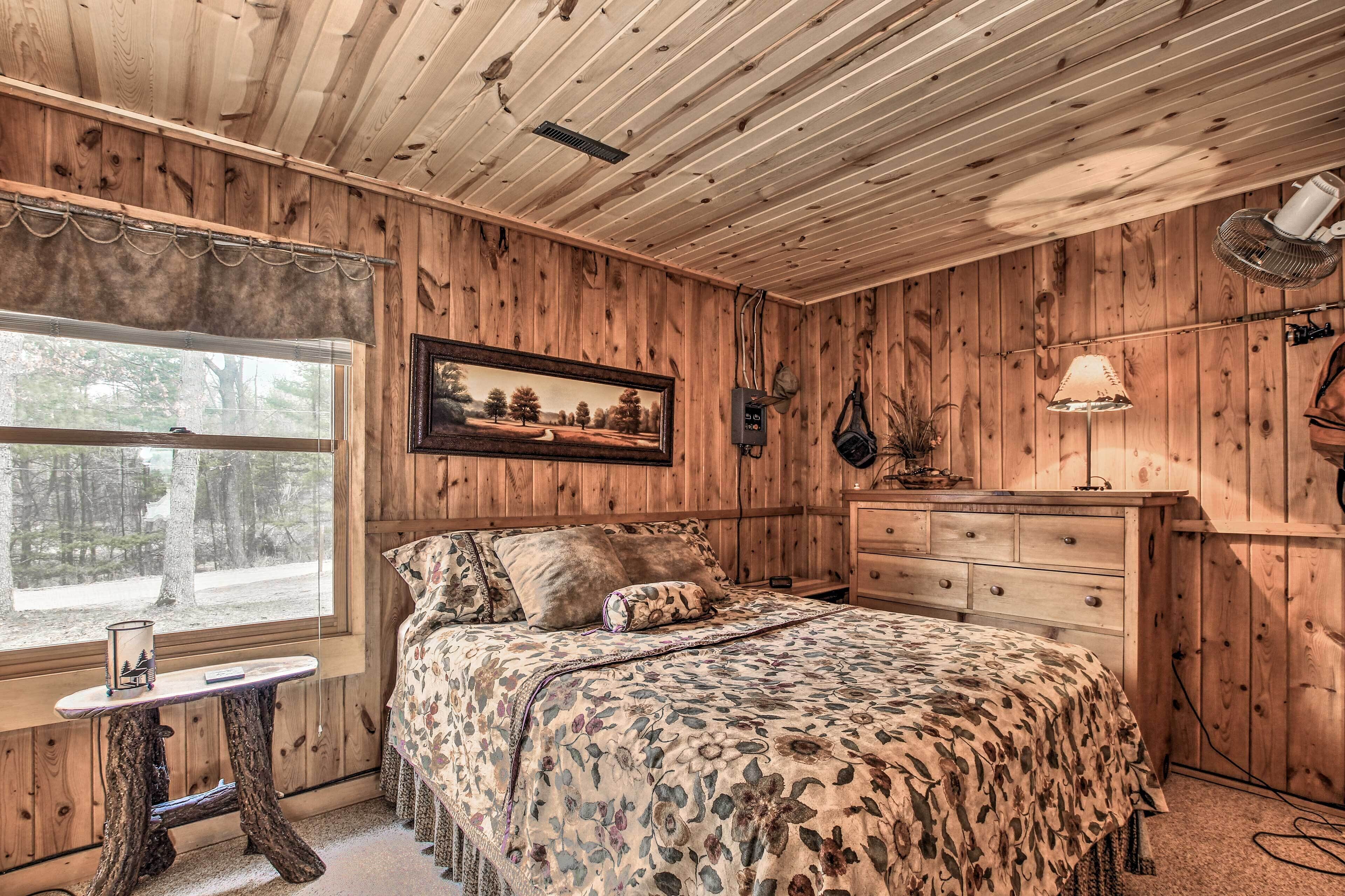 A wooden cabin bedroom with a bed, dresser, and a window with a view of trees.
