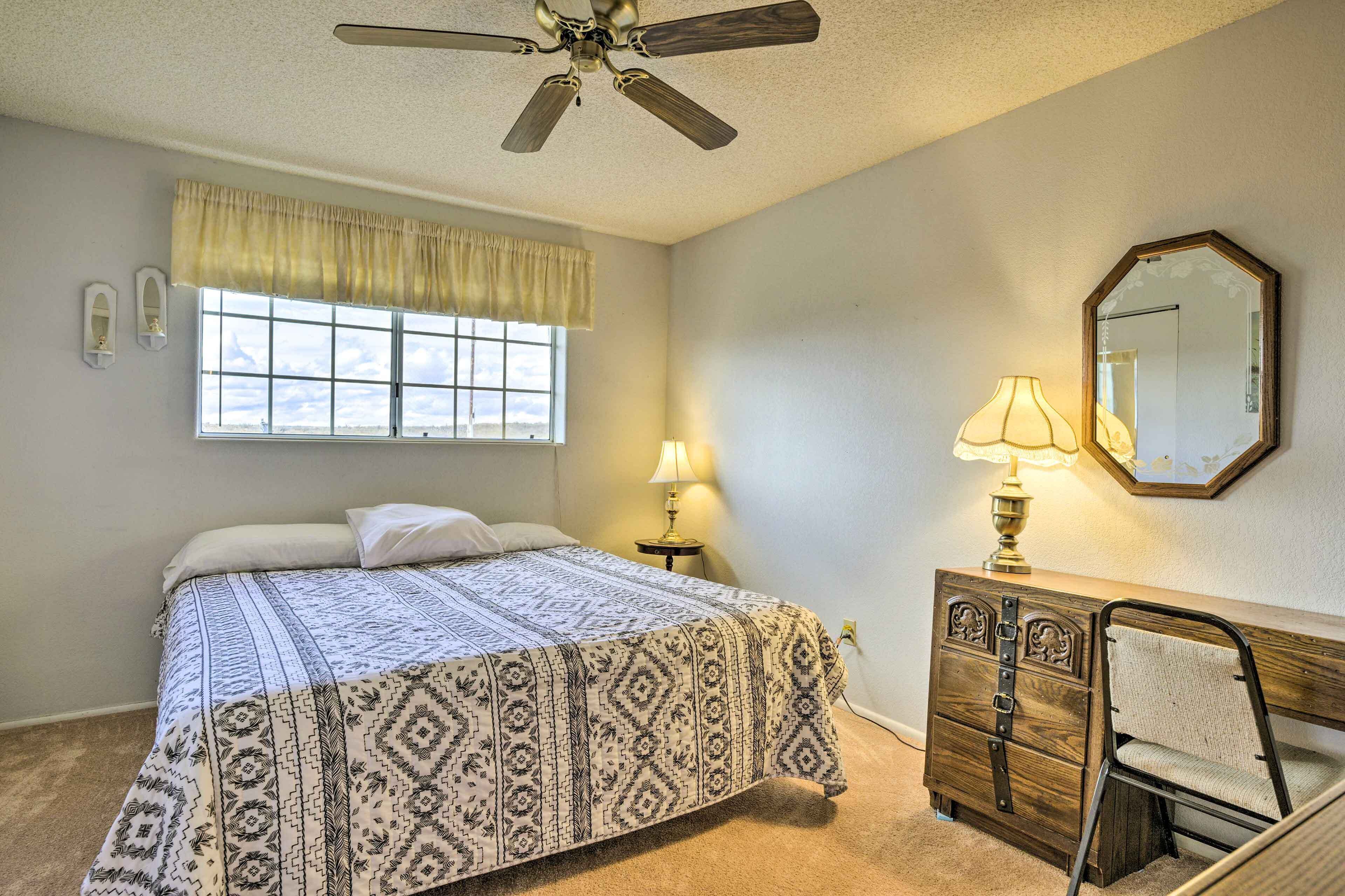 A bedroom with a bed, a desk, a chair, a ceiling fan, a window with curtains, a lamp, and a mirror.