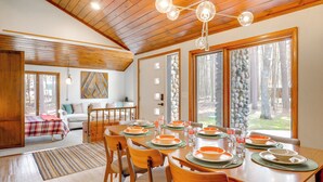 A dining area with a wooden table set for four, surrounded by chairs, with a view of trees through large windows. - Game Room! Mid-century Escape in the Northwoods (Woodruff)