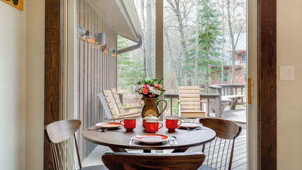 A dining table with chairs, a pitcher of flowers, and a view of a deck and trees. - Game Room! Mid-century Escape in the Northwoods (Woodruff)