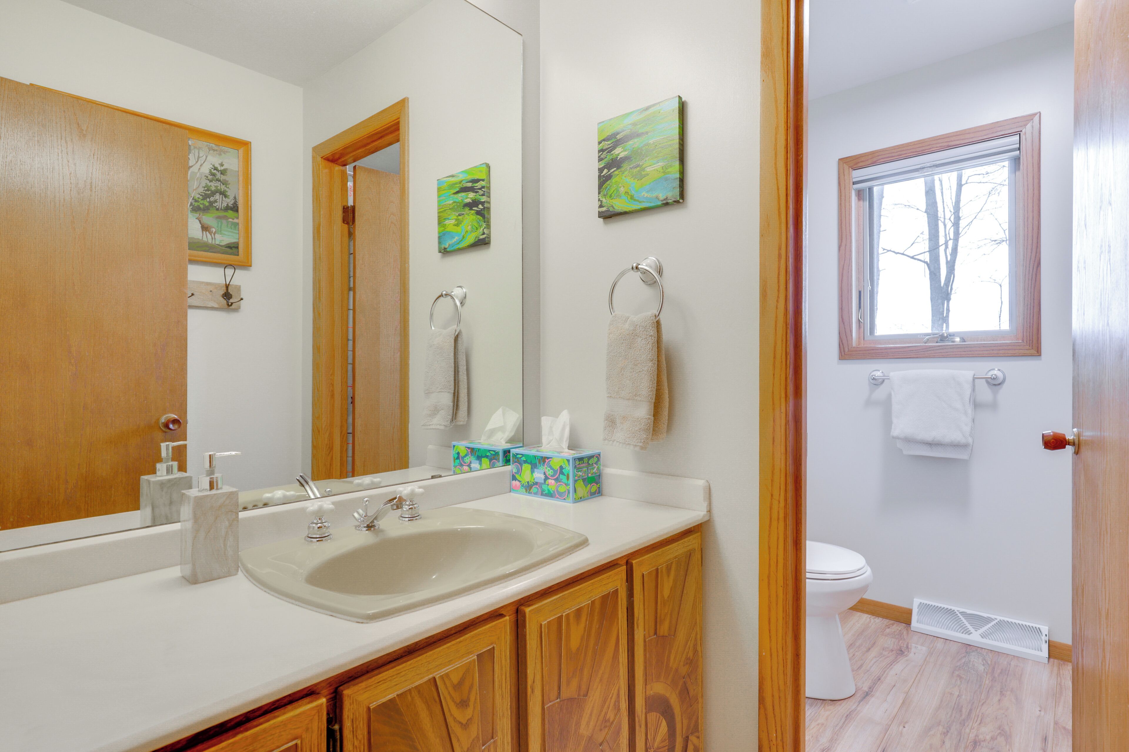 A bathroom with a sink, mirror, wooden cabinets, a toilet, and a window.