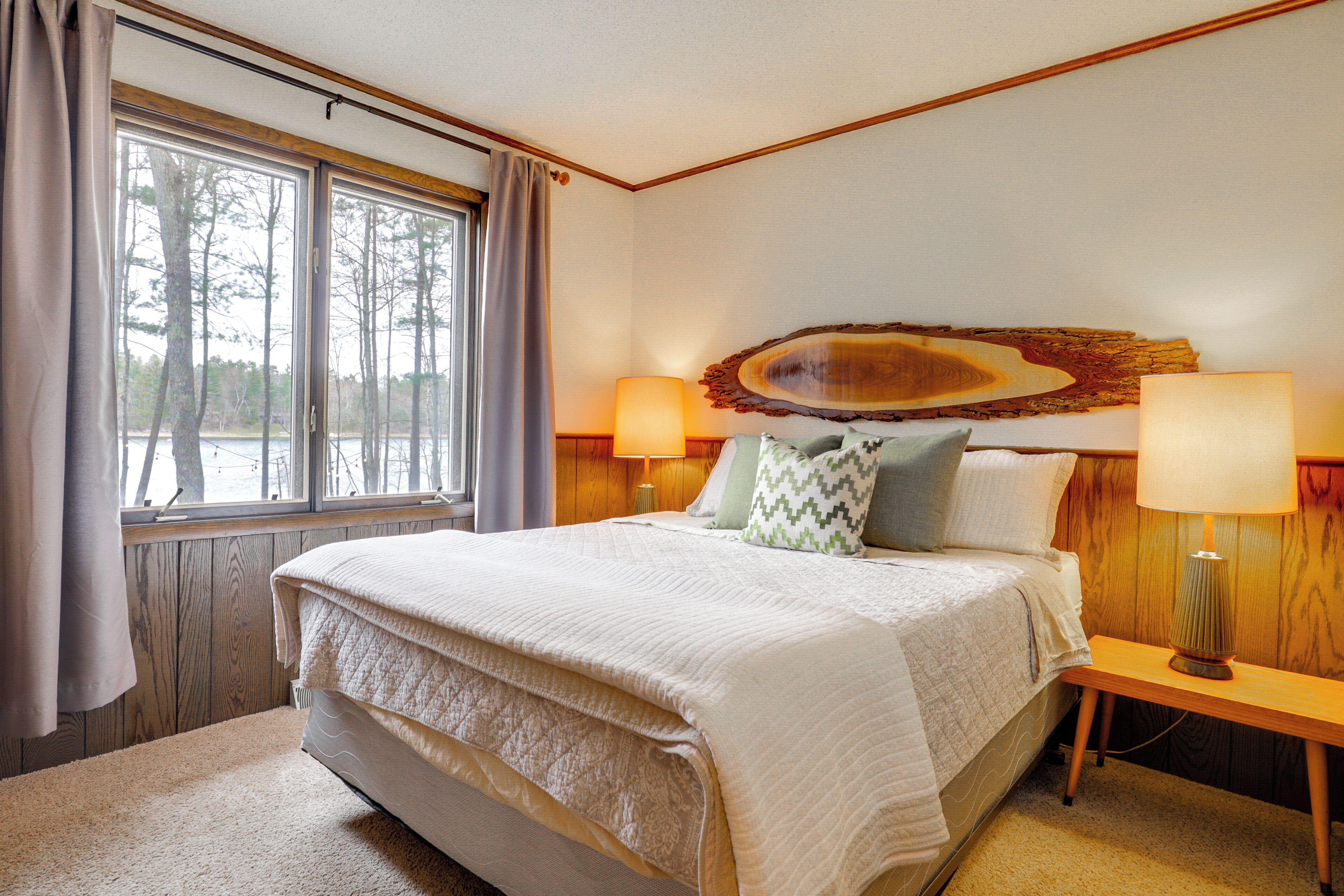 A bedroom with a large bed, two bedside lamps, a window with curtains, and a wooden headboard.