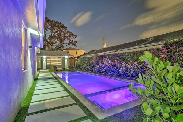 A swimming pool with a purple-lit edge, surrounded by a garden with purple flowers and green foliage.