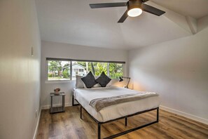 A bedroom with a bed, a window, a ceiling fan, and a small table with a lamp.