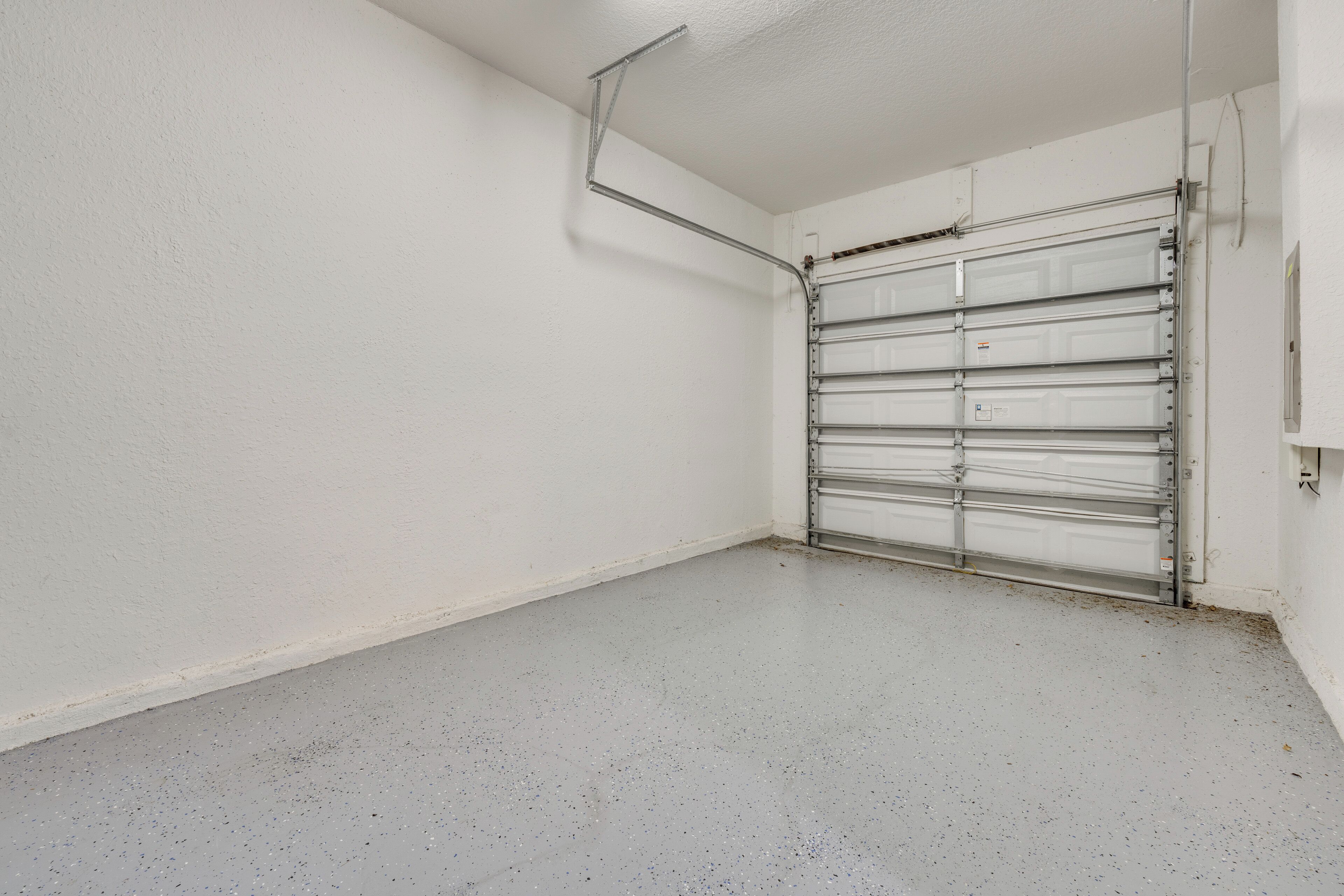 Un garage propre et vide, avec des murs blancs et un sol gris, doté d’une porte de garage en métal et d’un luminaire fixé au plafond.