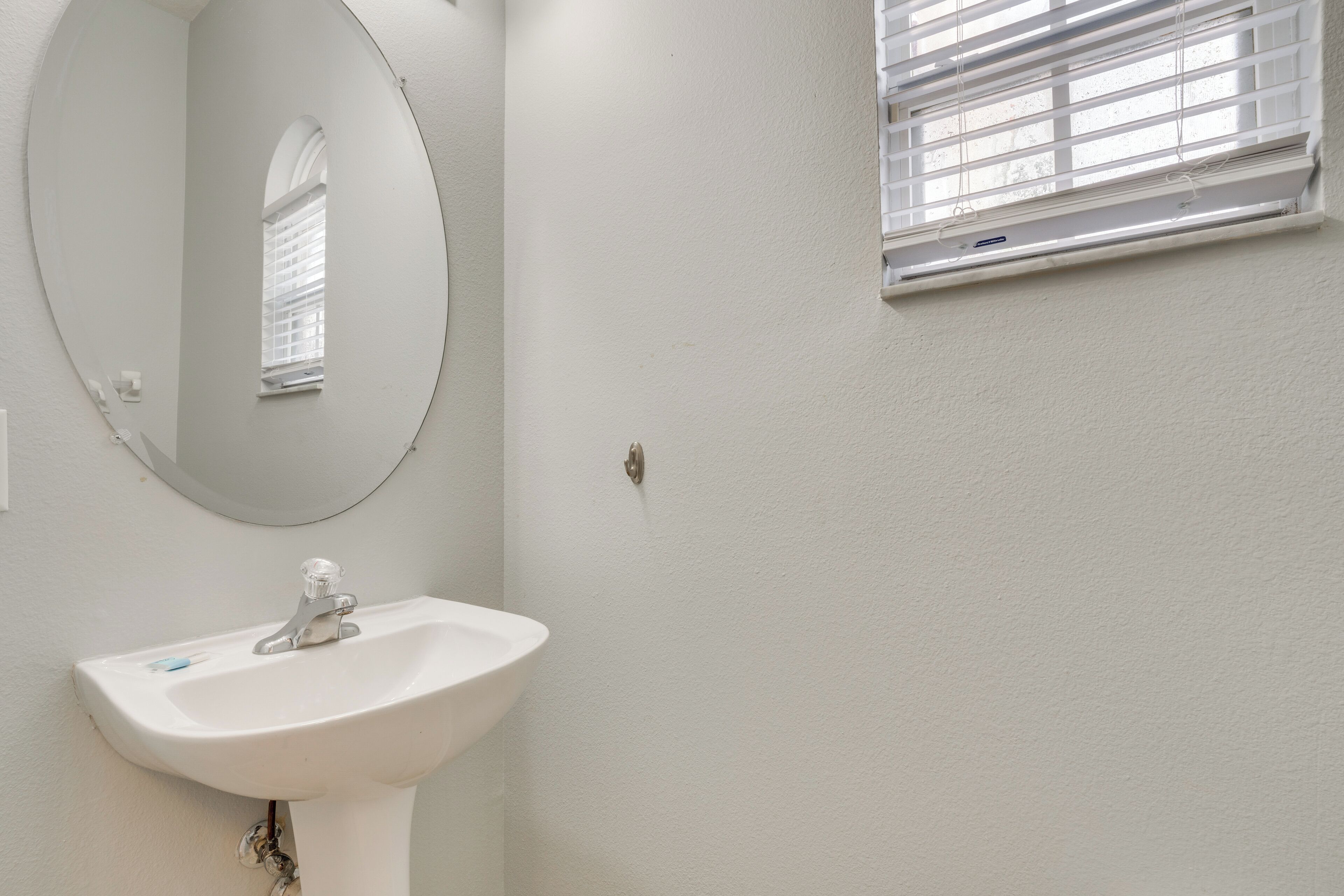 Une salle de bain avec un lavabo blanc, un miroir rond et une fenêtre avec des stores.