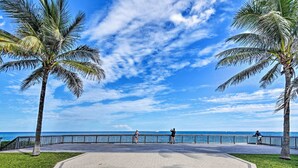 A paved walkway with palm trees, a railing, and a view of the ocean. - 1 Block to Deerfield Beach: Studio w/ Fire Pit! (Deerfield Beach)