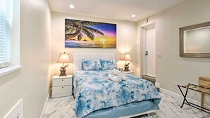 A bedroom with a bed, two bedside lamps, a nightstand, a wooden bench, and a large painting of a sunset over the ocean. - 1 Block to Deerfield Beach: Studio w/ Fire Pit! (Deerfield Beach)