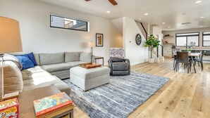 A spacious living area with a sofa, ottoman, and a dining table with chairs. A ceiling fan and a large window are visible. - Sleek Fraser Townhome < 8 Mi to Winter Park! (Fraser)