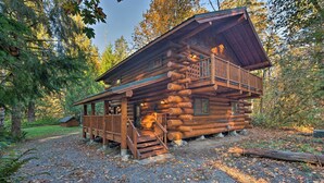 A log cabin with a balcony, a porch, and a small shed in a forested area. - Riverfront Gold Bar Log Cabin Near Stevens Pass! (Gold Bar)