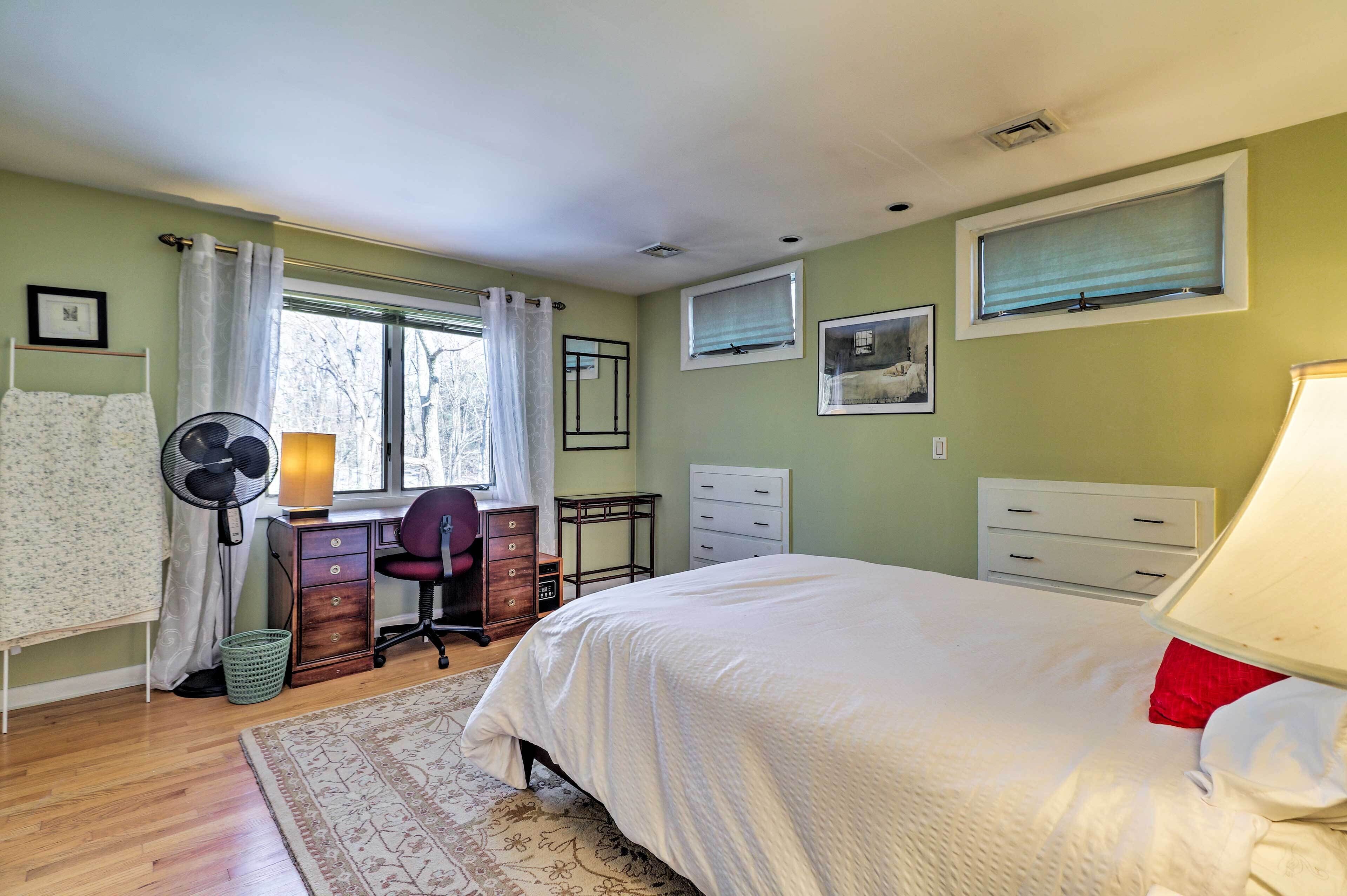 A bedroom with a bed, a desk with a chair, a fan, a window with curtains, and a dresser.
