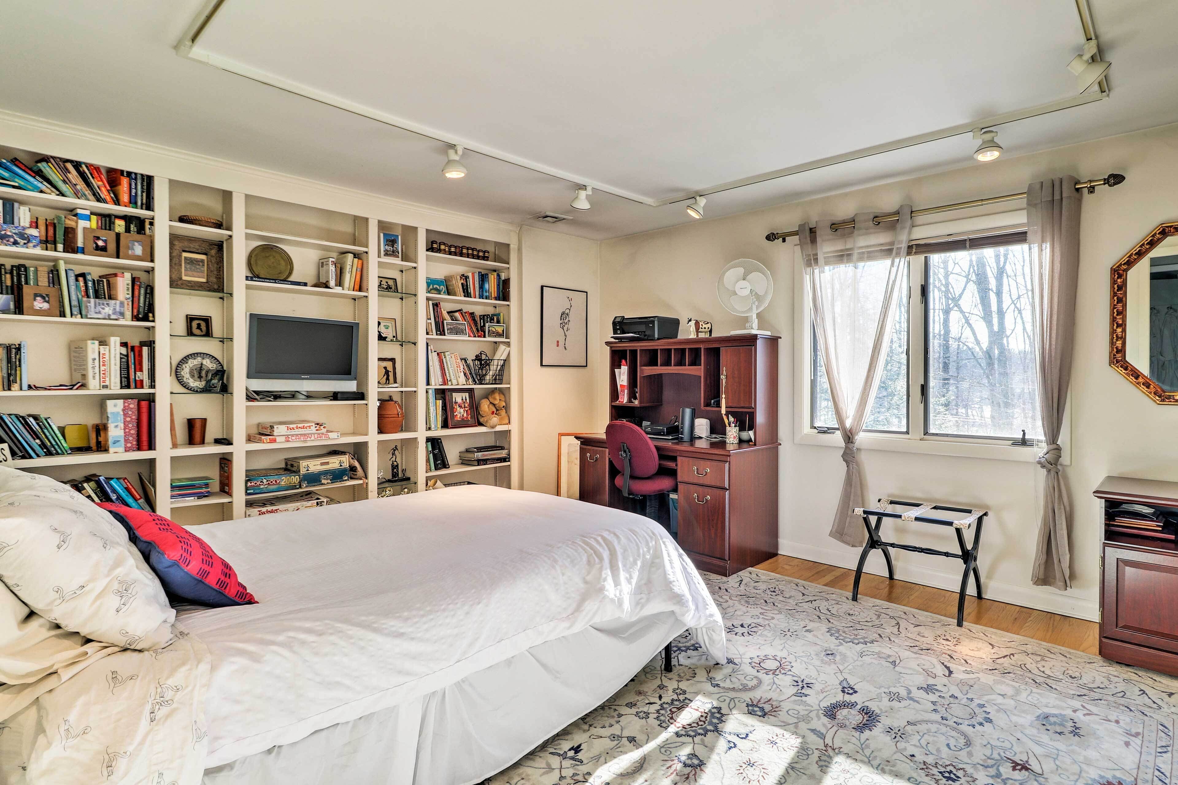 A bedroom with a large bed, a desk, a bookshelf, a television, and a window with curtains.